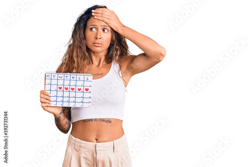 Young hispanic woman with tattoo holding heart calendar stressed and frustrated with hand on head, surprised and angry face