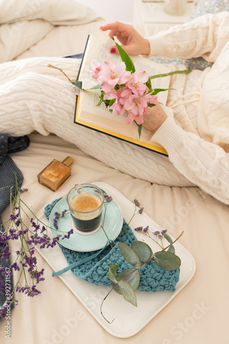 Woman reading a book on bed next to her coffee breakfast. She is holding a pretty bouquet of flowers with her left hand.