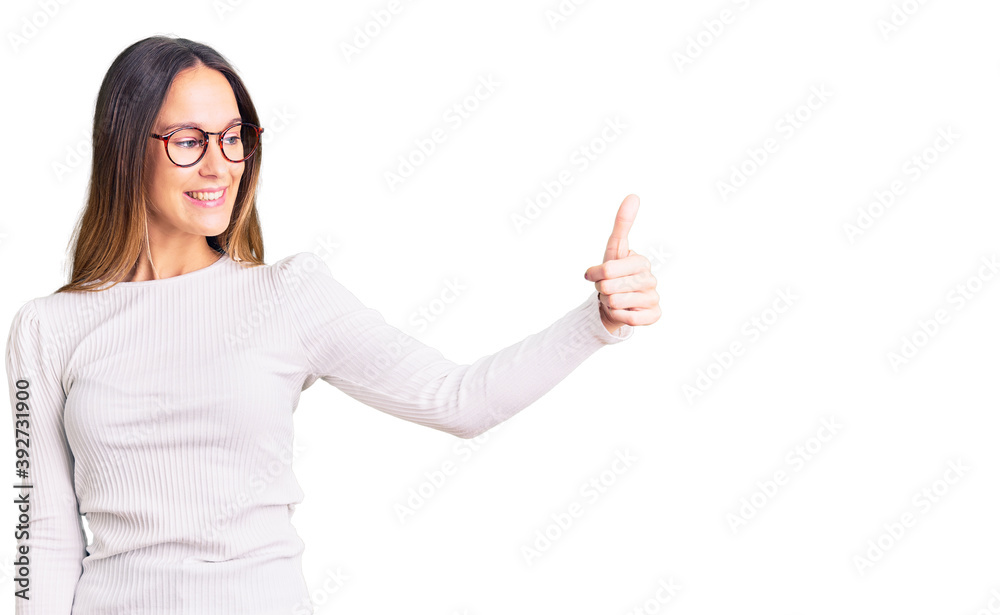 Beautiful brunette young woman wearing casual white sweater and glasses looking proud, smiling doing thumbs up gesture to the side Beautiful brunette young woman wearing casual white sweater and glasses looking proud, smiling doing thumbs up gesture to the side