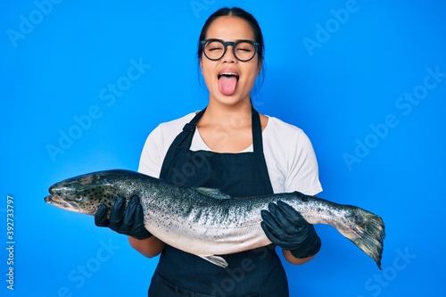 Young beautiful asian girl fishmonger selling fresh raw salmon sticking tongue out happy with funny expression.