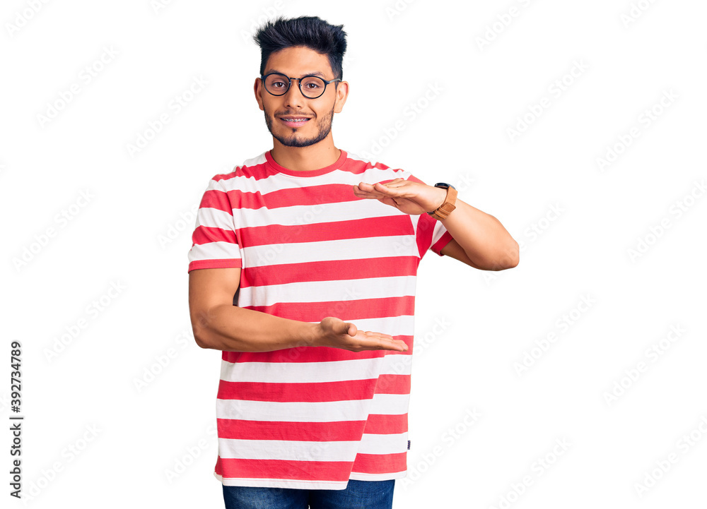 Handsome latin american young man wearing casual clothes and glasses gesturing with hands showing big and large size sign, measure symbol. smiling looking at the camera. measuring concept. Handsome latin american young man wearing casual clothes and glasses gesturing with hands showing big and large size sign, measure symbol. smiling looking at the camera. measuring concept.