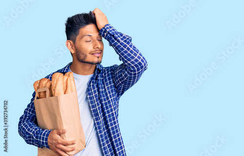 Handsome latin american young man holding paper bag with bread smiling confident touching hair with hand up gesture, posing attractive and fashionable