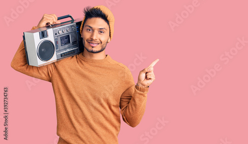 Handsome latin american young man holding boombox, listening to music cheerful with a smile on face pointing with hand and finger up to the side with happy and natural expression