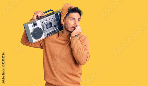Handsome latin american young man holding boombox, listening to music looking stressed and nervous with hands on mouth biting nails. anxiety problem.