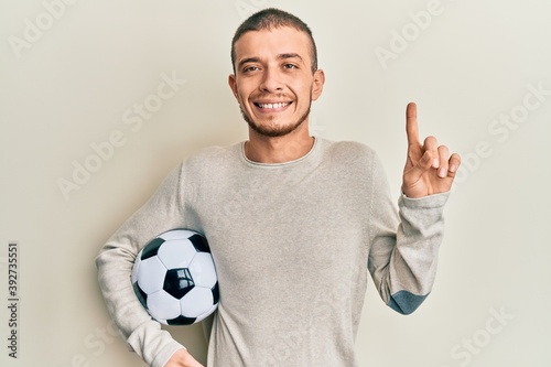 Young hispanic man holding soccer ball smiling happy pointing with hand and finger to the side © Krakenimages.com