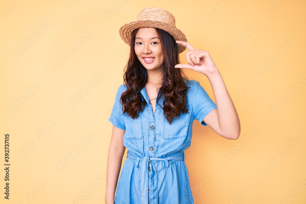 Young beautiful chinese girl wearing summer hat smiling and confident gesturing with hand doing small size sign with fingers looking and the camera. measure concept. Young beautiful chinese girl wearing summer hat smiling and confident gesturing with hand doing small size sign with fingers looking and the camera. measure concept.