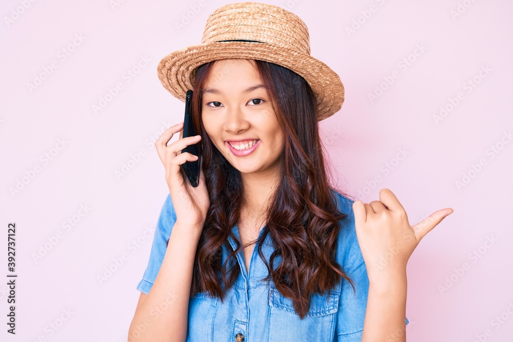 Young beautiful chinese girl wearing summer hat having conversation talking on the smartphone pointing thumb up to the side smiling happy with open mouth Young beautiful chinese girl wearing summer hat having conversation talking on the smartphone pointing thumb up to the side smiling happy with open mouth