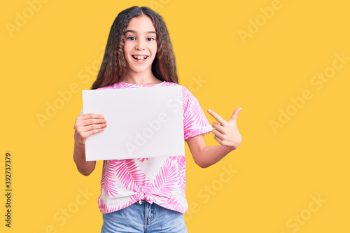 Cute hispanic child girl holding blank empty banner smiling happy pointing with hand and finger