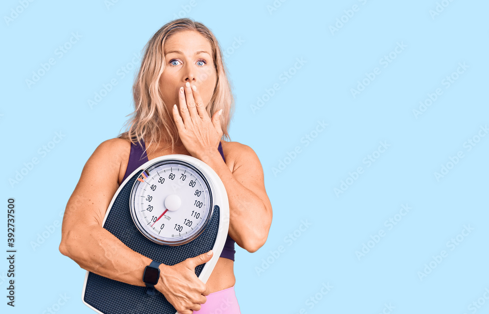 Middle age fit blonde woman wearing sports clothes holding weighing machine covering mouth with hand, shocked and afraid for mistake. surprised expression Middle age fit blonde woman wearing sports clothes holding weighing machine covering mouth with hand, shocked and afraid for mistake. surprised expression