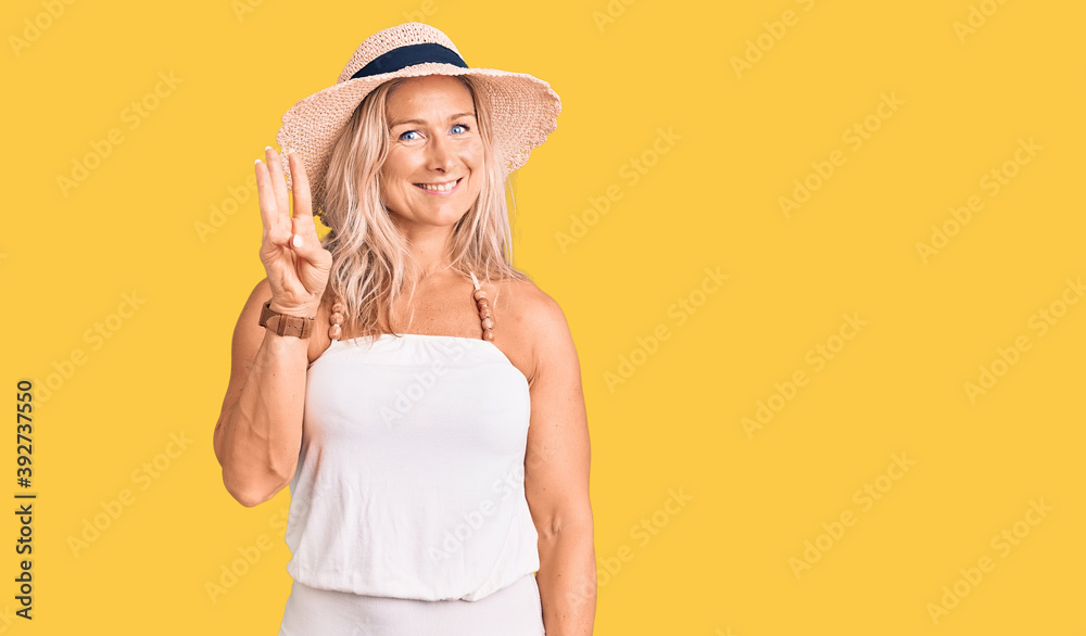 Middle age fit blonde woman wearing summer hat showing and pointing up with fingers number three while smiling confident and happy. Middle age fit blonde woman wearing summer hat showing and pointing up with fingers number three while smiling confident and happy.