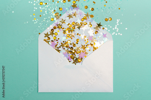 Sparkling confetti pouring out of white envelope on mint background. View from above. Flat lay. Holiday, congratulation or good news concept.