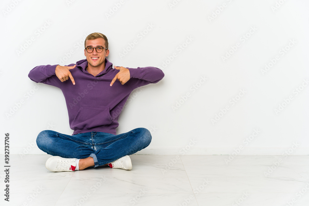 Young caucasian man sitting on the floor points down with fingers, positive feeling.