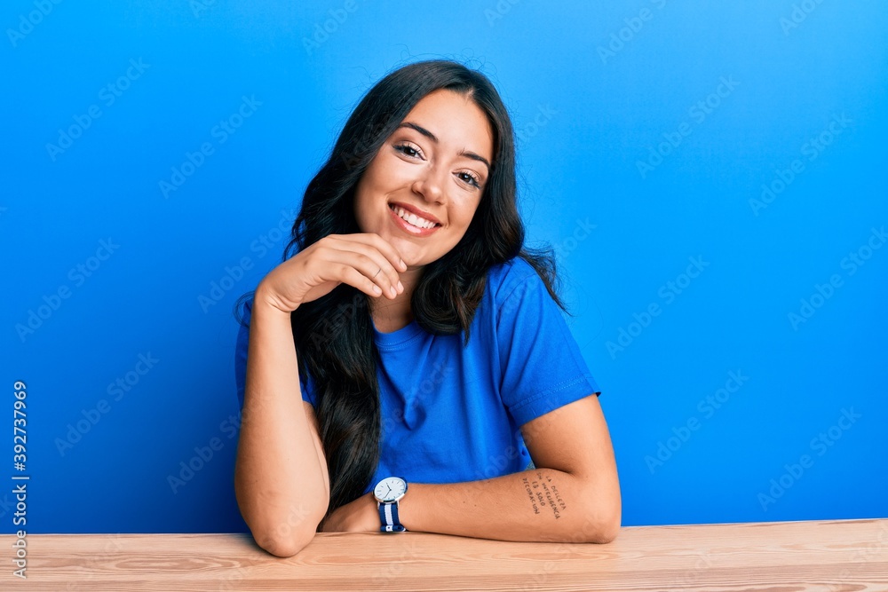 Beautiful brunette young woman wearing casual clothes sitting on the table smiling looking confident at the camera with crossed arms and hand on chin. thinking positive. Beautiful brunette young woman wearing casual clothes sitting on the table smiling looking confident at the camera with crossed arms and hand on chin. thinking positive.