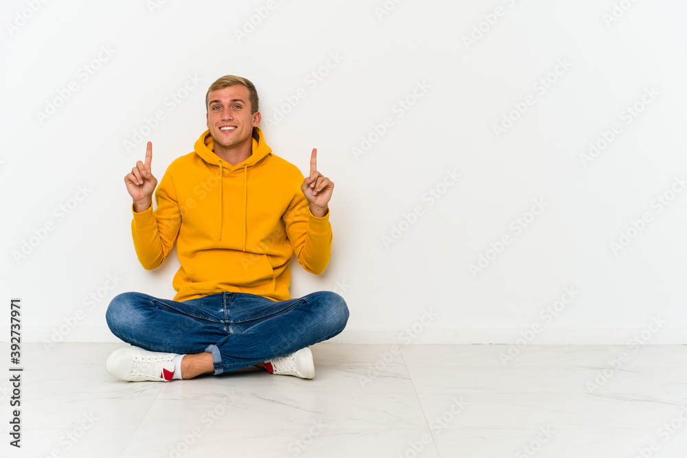 Young caucasian man sitting on the floor indicates with both fore fingers up showing a blank space. Young caucasian man sitting on the floor indicates with both fore fingers up showing a blank space.