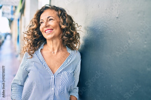 Middle age hispanic woman smiling happy leaning on the wall at the city.