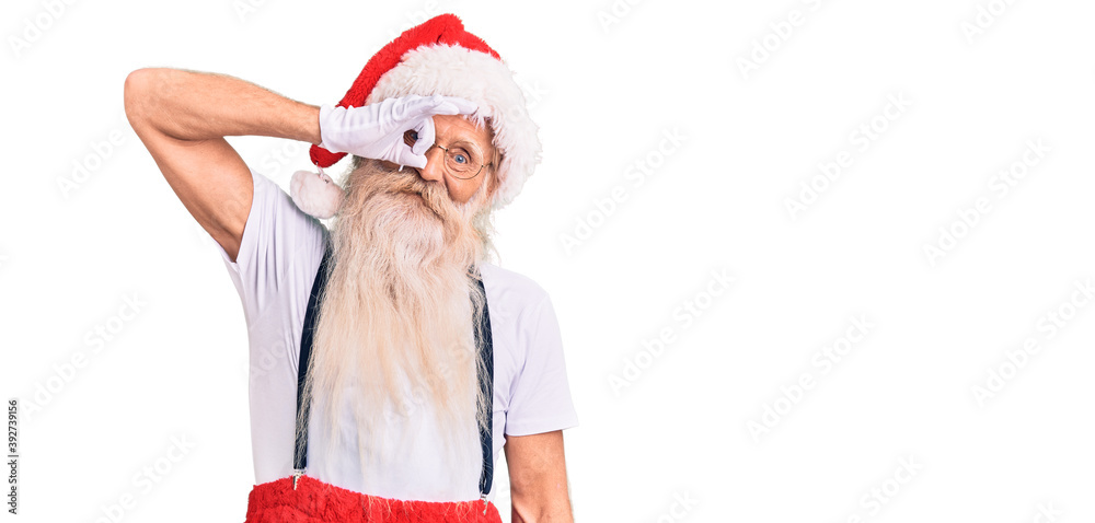 Obraz premium Old senior man with grey hair and long beard wearing santa claus costume with suspenders smiling happy doing ok sign with hand on eye looking through fingers