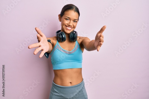 Beautiful hispanic woman wearing sportswear and headphones looking at the camera smiling with open arms for hug. cheerful expression embracing happiness.