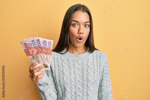 Beautiful hispanic woman holding 100 hong kong dollars banknotes scared and amazed with open mouth for surprise, disbelief face