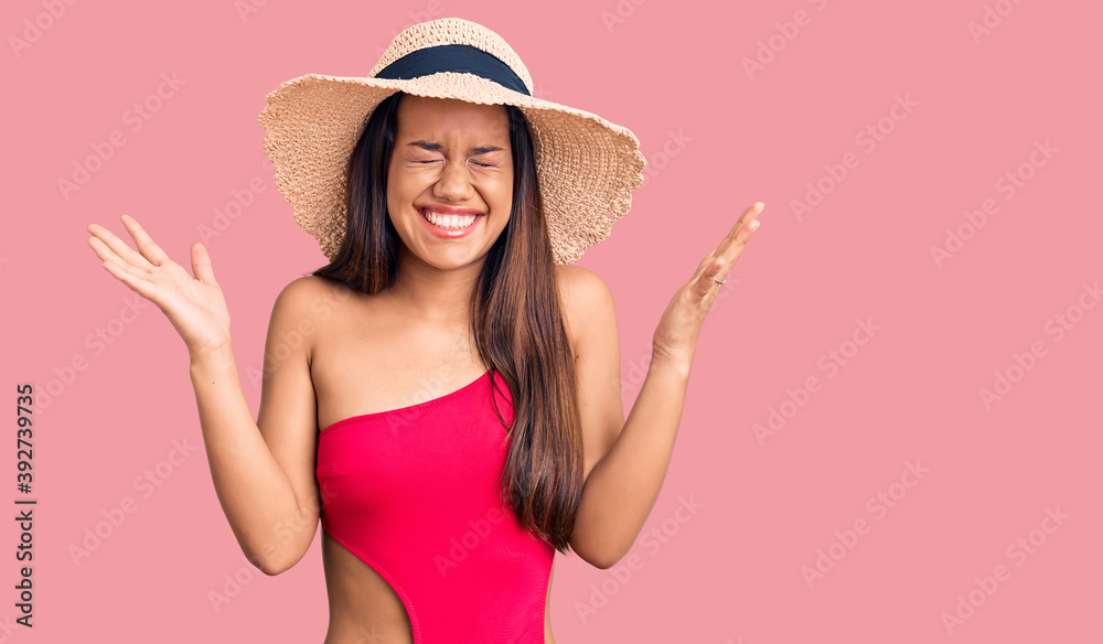 Young beautiful latin girl wearing swimwear and summer hat celebrating mad and crazy for success with arms raised and closed eyes screaming excited. winner concept