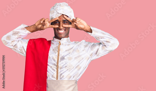 African handsome man wearing tradition sherwani saree clothes doing peace symbol with fingers over face, smiling cheerful showing victory