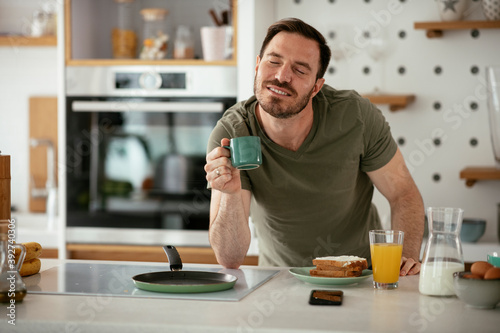 Handsome man preparing breakfast at home. Young man drinking coffee while preparing sandwich.