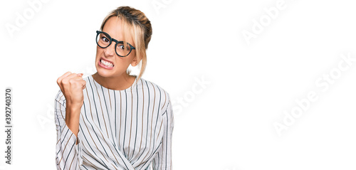 Beautiful blonde woman wearing business shirt and glasses angry and mad raising fist frustrated and furious while shouting with anger. rage and aggressive concept.