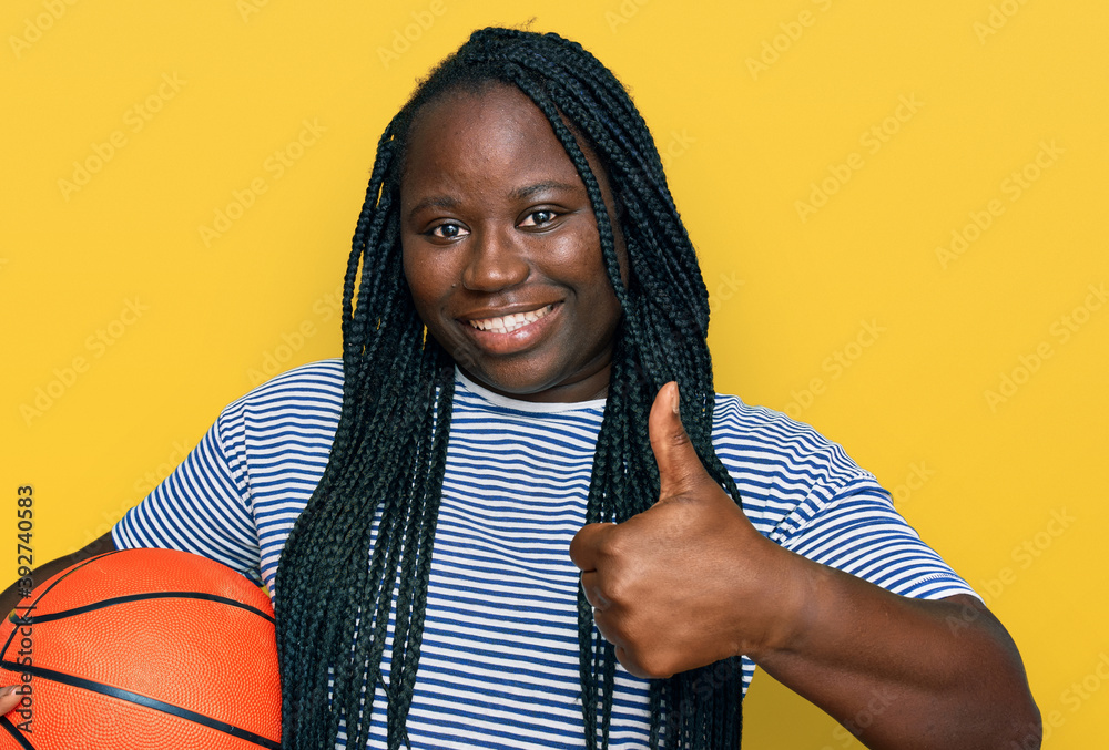 Young black woman with braids holding basketball ball smiling happy and positive, thumb up doing excellent and approval sign Young black woman with braids holding basketball ball smiling happy and positive, thumb up doing excellent and approval sign