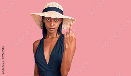 Young african american woman wearing swimsuit and summer hat showing middle finger, impolite and rude fuck off expression