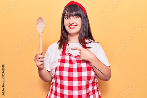 Young plus size woman wearing apron holding wooden spoon smiling happy pointing with hand and finger
