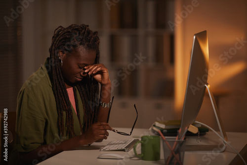 Side view portrait of exhausted African-American woman rubbing eyes while working late at night in dark office, copy space photo