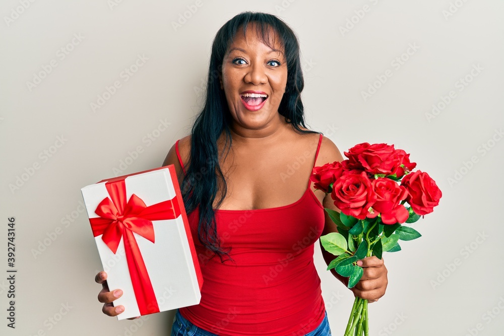 Middle age african american woman holding anniversary present and bouquet of flowers smiling and laughing hard out loud because funny crazy joke. Middle age african american woman holding anniversary present and bouquet of flowers smiling and laughing hard out loud because funny crazy joke.