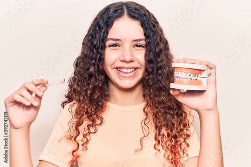 Adorable latin teenager smiling happy. Standing with smile on face holding denture with bracket and aligner over isolated white background photo