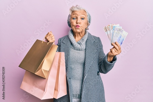 Senior grey-haired woman holding shopping bags and swedish krona banknotes puffing cheeks with funny face. mouth inflated with air, catching air.