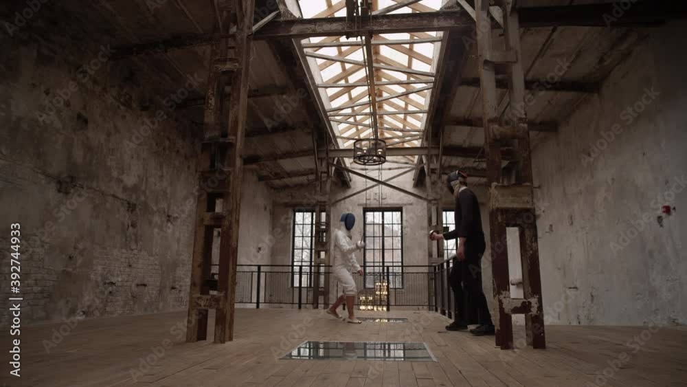 Two fencers in white and black suits and masks compete with sabers in the old loft room where the dark side wins. The concept of the struggle between good and evil. Slow motion Two fencers in white and black suits and masks compete with sabers in the old loft room where the dark side wins. The concept of the struggle between good and evil. Slow motion