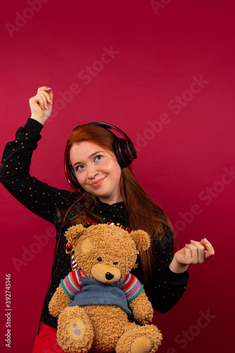 A cute girl with a bear, listens to music in big headphones and dances. Photo on a pink background. Spending time concept. photo