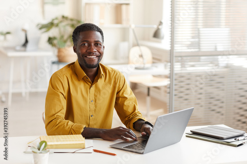 Portrait of smiling African-American man using laptop and looking at camera while enjoying work in minimal office interior, copy space