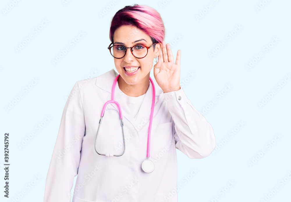 Young beautiful woman with pink hair wearing doctor uniform smiling with hand over ear listening an hearing to rumor or gossip. deafness concept. Young beautiful woman with pink hair wearing doctor uniform smiling with hand over ear listening an hearing to rumor or gossip. deafness concept.