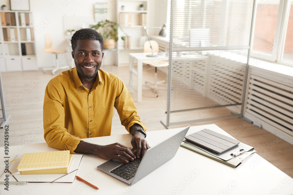 High angle portrait of smiling African-American man using laptop and looking at camera while enjoying work in minimal office interior, copy space - obrazy, fototapety, plakaty 