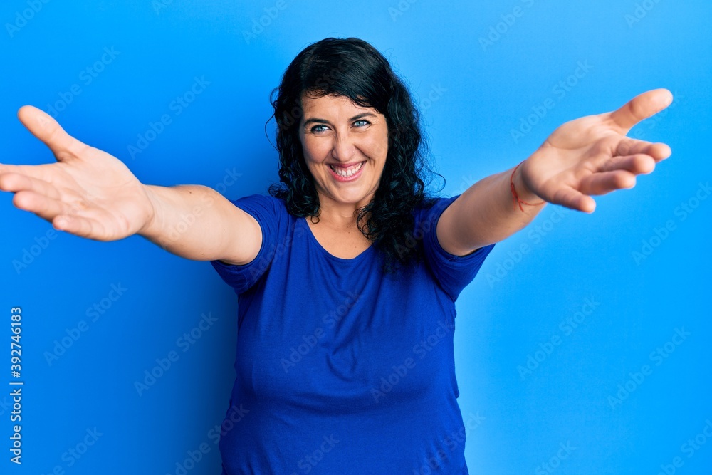 Plus size brunette woman wearing casual blue shirt looking at the camera smiling with open arms for hug. cheerful expression embracing happiness. Plus size brunette woman wearing casual blue shirt looking at the camera smiling with open arms for hug. cheerful expression embracing happiness.