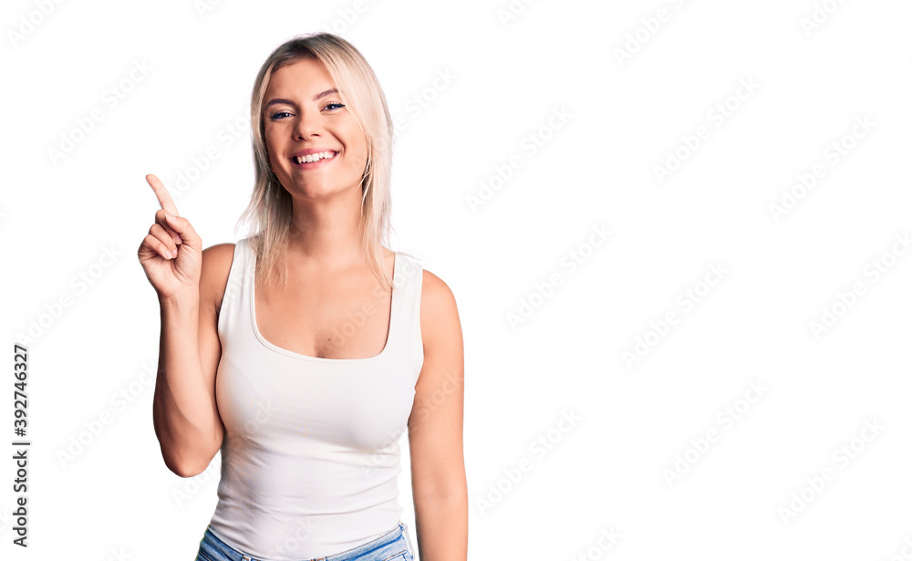 Young beautiful blonde woman wearing casual sleeveless t-shirt showing and pointing up with finger number one while smiling confident and happy. Young beautiful blonde woman wearing casual sleeveless t-shirt showing and pointing up with finger number one while smiling confident and happy.