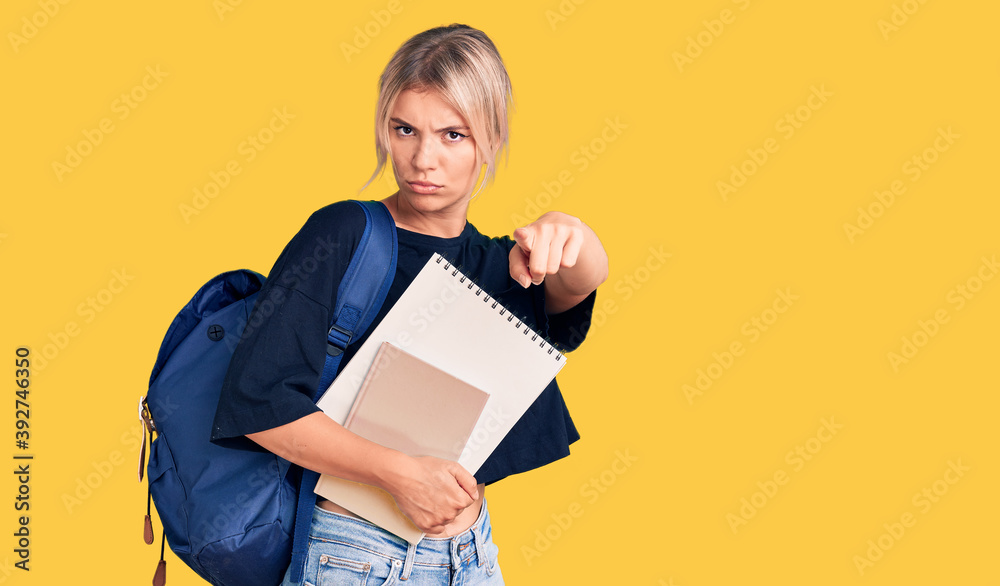Young beautiful blonde woman wearing student backpack holding notebook pointing with finger to the camera and to you, confident gesture looking serious Young beautiful blonde woman wearing student backpack holding notebook pointing with finger to the camera and to you, confident gesture looking serious