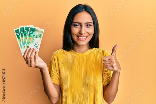 Young brunette woman holding 50 israel shekels banknotes smiling happy and positive, thumb up doing excellent and approval sign