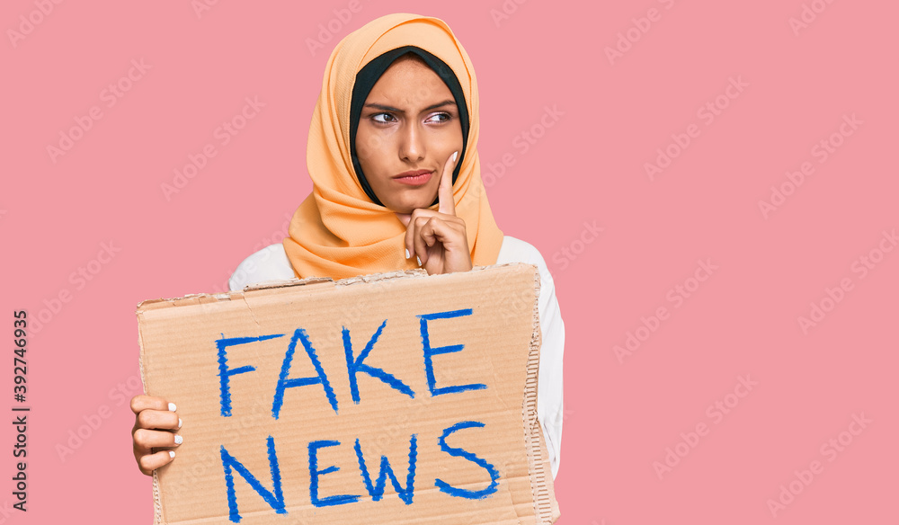 Young brunette arab woman holding fake news banner serious face thinking about question with hand on chin, thoughtful about confusing idea Young brunette arab woman holding fake news banner serious face thinking about question with hand on chin, thoughtful about confusing idea