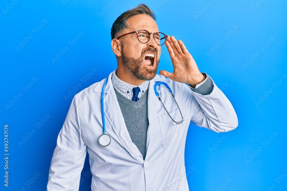 Handsome middle age man wearing doctor uniform and stethoscope shouting and screaming loud to side with hand on mouth. communication concept. Handsome middle age man wearing doctor uniform and stethoscope shouting and screaming loud to side with hand on mouth. communication concept.