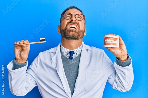 Handsome middle age dentist man holding denture and toothbrush angry and mad screaming frustrated and furious  shouting with anger looking up.