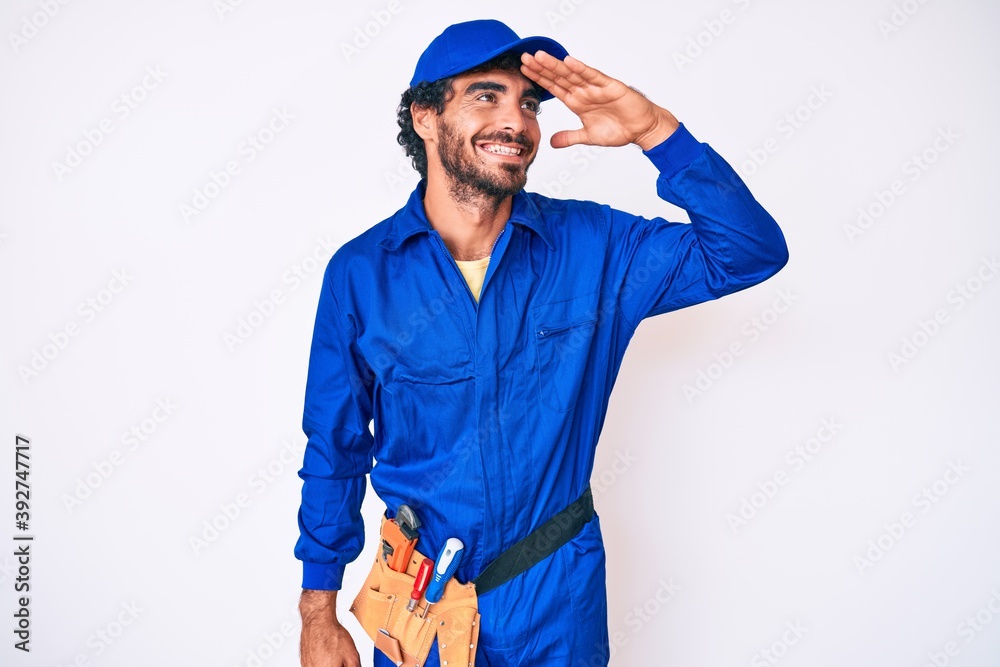 Handsome young man with curly hair and bear weaing handyman uniform very happy and smiling looking far away with hand over head. searching concept.