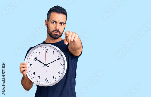 Young hispanic man holding big clock pointing with finger to the camera and to you, confident gesture looking serious