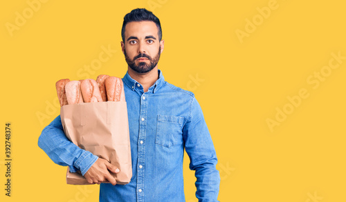 Young hispanic man holding bag with bread thinking attitude and sober expression looking self confident photo