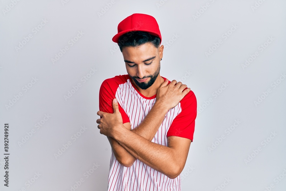 Obraz premium Young man with beard wearing baseball uniform hugging oneself happy and positive, smiling confident. self love and self care
