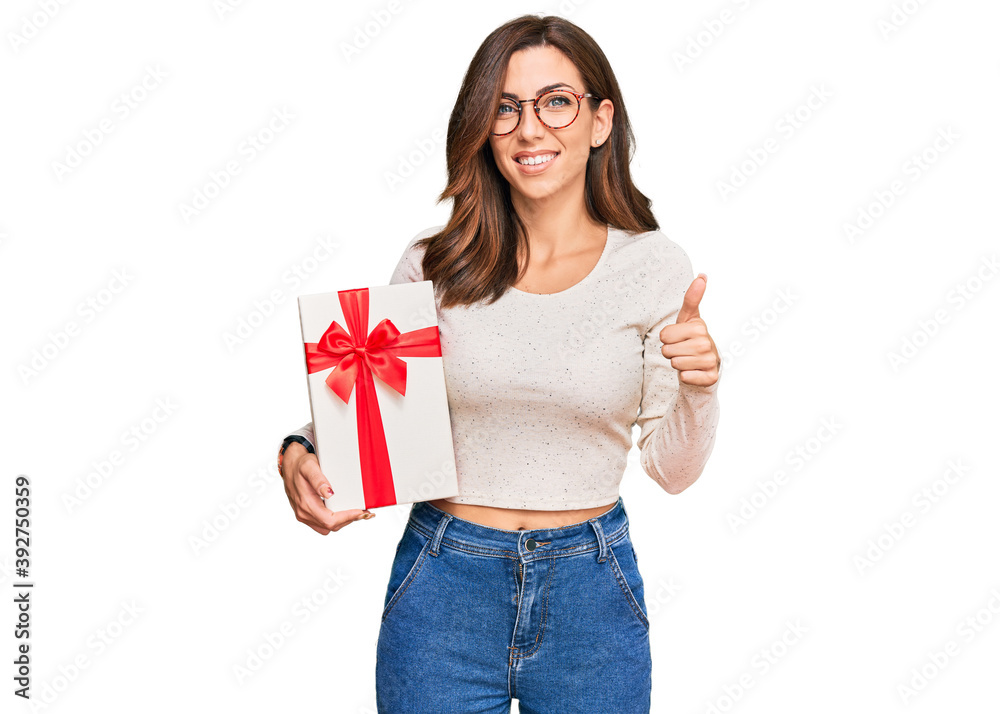 Young brunette woman holding gift smiling happy and positive, thumb up doing excellent and approval sign Young brunette woman holding gift smiling happy and positive, thumb up doing excellent and approval sign